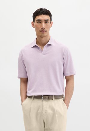 Man met kort donker haar die een lichtpaarse polo met korte mouwen draagt, ingestopt in een beige broek met een bruine riem, handen in de zakken.
