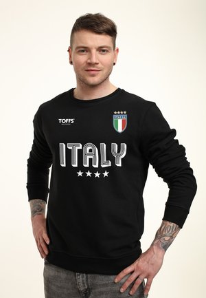 Młody mężczyzna z krótkimi włosami i tatuażami, ubraną w czarną bluzę z napisem "ITALY", czterema gwiazdkami i herbem reprezentacji Włoch w piłce nożnej.