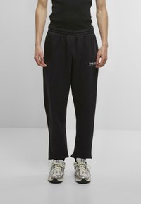 Black jogger pants with elastic waistband, featuring a straight cut and printed logo on the right side. Paired with silver and black sneakers.