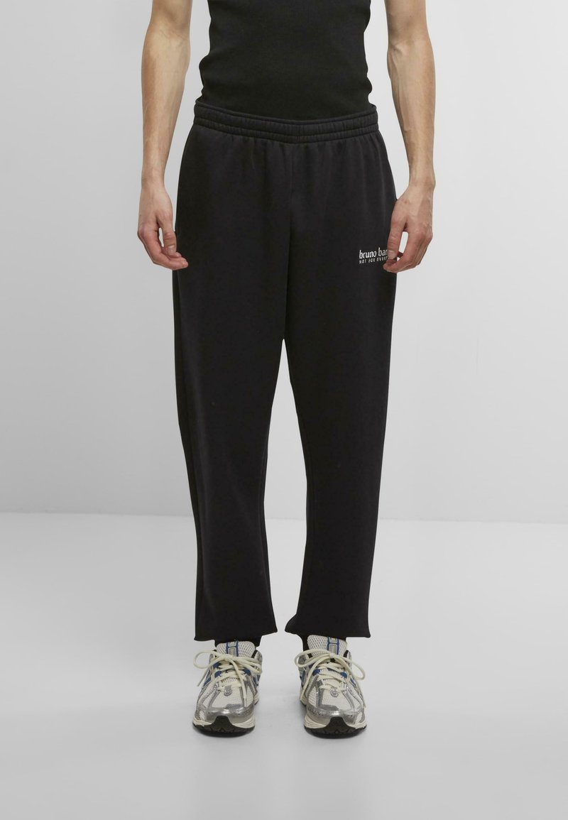 Black jogger pants with elastic waistband, featuring a straight cut and printed logo on the right side. Paired with silver and black sneakers.