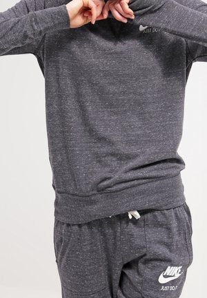 Persoon die een donkergrijs Nike-trui en broek draagt en met beide handen de kraag van de trui aanpast.