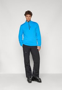 Πετσετάκι μπλε με μακριά μανίκια και μισό-φερμουάρ στο γιακά, με λογότυπο. Μαύρο παντελόνι με λεπτομέρεια στους πλευρικούς μ pockets. Και τα δύο ρούχα είναι φτιαγμένα από μαλακό, ελαφρύ ύφασμα.