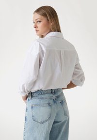 Camicia bianca con un sottile motivo a pois, vestibilità rilassata e maniche lunghe. Abbinata a jeans in denim a vita alta di color azzurro chiaro.