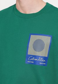 Zelené bavlněné tričko s kulatým výstřihem, s modrou grafickou nášivkou a vertikálním pruhovaným vzorem, zobrazující kruhový tmavě šedý prvek.