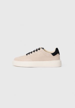 Basket basse en daim beige avec lacets noirs et languette au talon, semelle blanche épaisse, vue de profil sur fond clair.