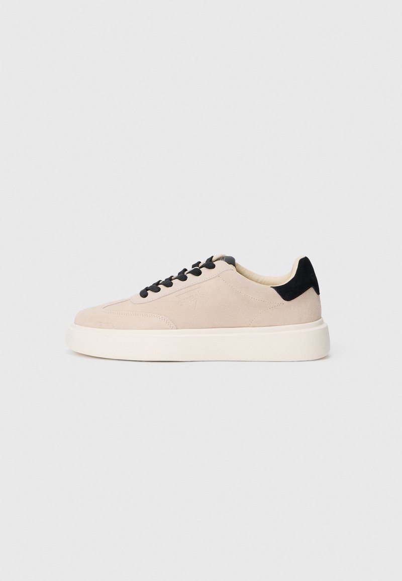 Basket basse en daim beige avec lacets noirs et languette au talon, semelle blanche épaisse, vue de profil sur fond clair.