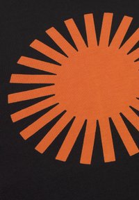 Čierna tkanina s veľkým oranžovým vzorom slnečného žiarenia, ktorý obsahuje striedavé lúče a medzery, čím vytvára výrazný vizuálny kontrast.