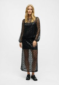 Schwarzes Pailletten-Kleid mit langen, transparenten Ärmeln und einem knielangen Rock, der eine strukturierte Überlage und eine enganliegende Innenschicht aufweist.