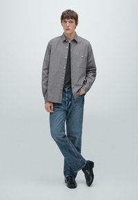 Grijze button-up shirt met een gestructureerd patroon, gedragen over een donker t-shirt. Gepaard met blauwe jeans en zwarte schoenen. Staande pose op een neutrale achtergrond.