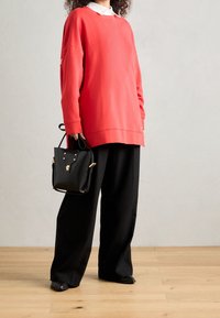 Röd överdimensionerad sweatshirts, vit krage, parat med lösa svarta byxor och en strukturerad svart handväska med gulddetaljer. Trägolvsbakgrund.