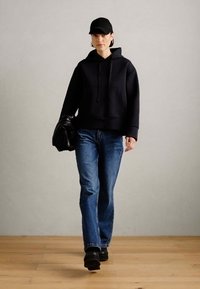 Black hooded sweatshirt with a kangaroo pocket, paired with blue straight-leg jeans and black footwear. Accompanied by a black cap and bag.