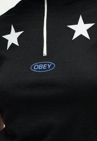 Μαύρο πουκάμισο με λευκό φερμουάρ, δύο λευκά αστέρια στο στήθος και μπλε οβάλ λογότυπο με τη λέξη «OBEY» στο κέντρο.