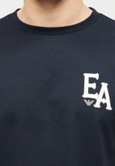 Κοντινό πλάνο σκούρου μπλε μπλουζάκι με λευκά γράμματα "EA" και μικρό λογότυπο στο στήθος, φορεμένο από άτομο που φαίνεται από τον λαιμό μέχρι το πάνω μέρος του στήθους.
