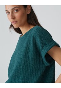 Teal gemêleerde top met een losse pasvorm, voorzien van een ronde halslijn en opgerolde mouwen. De stof heeft een opvallend, gekreukt patroon.