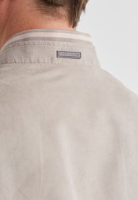 Hellbeige Wildlederjacke mit glatter Textur, ausgestattet mit einem gestreiften Kragen und einem Metallmarkenetikett auf der Rückseite.