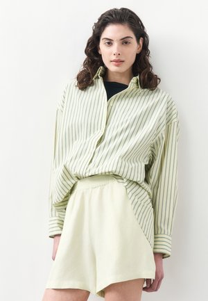 Femeie cu păr creț și închis la culoare, purtând o cămașă largă cu dungi verzi pal peste un top negru și pantaloni scurți crem cu talie elastică, pe un fundal alb.