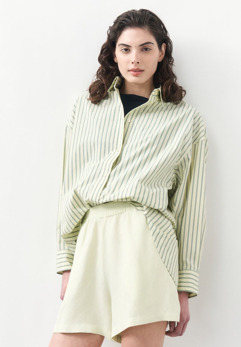 Femme aux cheveux bouclés foncés portant une chemise ample à rayures vert pâle sur un haut noir et un short à taille élastique de couleur crème, sur fond blanc.