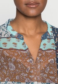 Blusa multi-pattern con colletto, blocchi di colore in teal, marrone e crema, con design floreali e paisley.