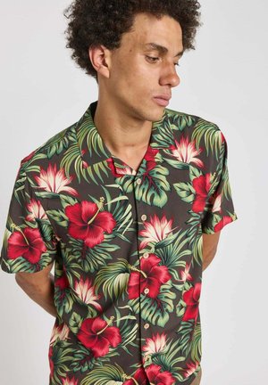Jeune homme aux cheveux bouclés portant une chemise noire à manches courtes ornée de motifs floraux tropicaux rouges et blancs.