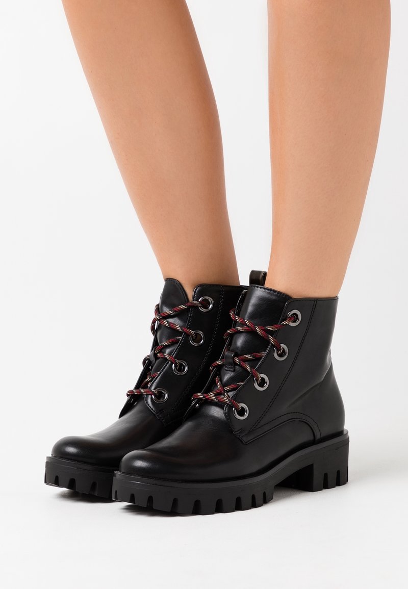 Stivaletti neri in pelle con suole spesse e lacci con accenti rossi indossati su gambe nude su uno sfondo bianco.