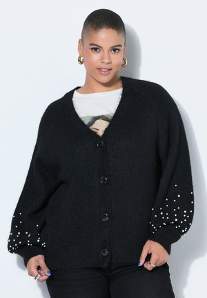 Person med kort hår iført en sort cardigan med knapper og perlebesætning på ærmerne samt en hvid grafisk T-shirt under.