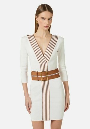 Donna che indossa un vestito bianco con dettagli a righe marroni a V sul collo e una larga cintura marrone con fibbia dorata, in piedi su uno sfondo bianco semplice.