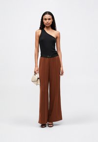 Top negru cu un umăr, asortat cu pantaloni maro cu talie înaltă și lățiți. Modelul ține o poșetă mică bej cu accente negre.