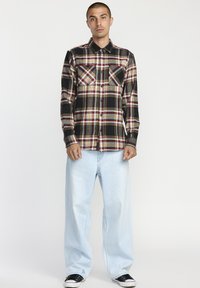 Geruit overhemd in zwart, rood en geel met twee borstzakken; gecombineerd met lichtblauwe baggy jeans en zwarte sneakers.