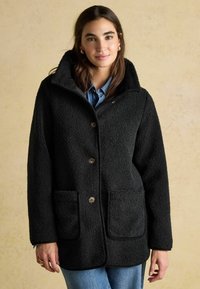 Mustavilla fleecetakki, jossa on teksturoitu pinta, suuri kaulus, nappikynnyksellä suljettava ja kaksi etutaskua. Päällä farkkupaita.