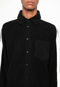 Čierna fleecová košeľa s golierom, s čiernymi gombíkmi, jednou náprsníkovou kapsou a bielymi bočnými pruhmi s čiernymi detailmi.