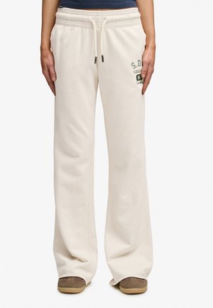 Person iført løse off-white joggingbukser med snøre i taljen og lille trykt logo på øverste venstre lår, kombineret med brune sko.