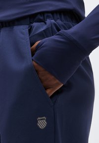 Χέρι στην τσέπη μπλε σκούρου αθλητικού παντελονιού με ελαστική μέση και λογότυπο K-Swiss στο μηρό, φορεμένο με αντίστοιχο μακρυμάνικο τοπ.