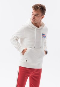 Sudadera blanca con un bolsillo delantero, presenta un gráfico de "OMG" en azul y naranja. Se lleva con pantalones de chándal rojos con franjas laterales. Textura de tela suave.