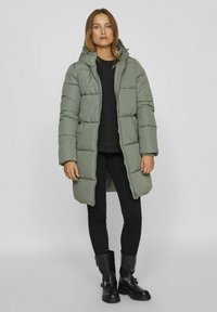 Cazadora acolchada verde con capucha, con un diseño cuadrado de acolchado y puños elásticos. Llevada sobre una camiseta negra y combinada con leggings y botas negras.