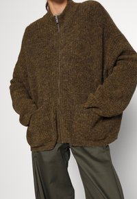 Brauner gerippter Strickcardigan mit hohem Kragen und Frontreißverschluss. Verfügt über zwei Seiten Taschen und einen lockeren Schnitt. Gemütliches, strukturiertes Material.