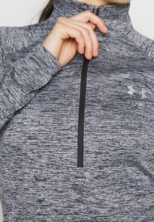 Hand hält schwarzen Reißverschluss an grauem, strukturiertem langärmligem Sportoberteil mit hohem Kragen und Logo auf der Brust.