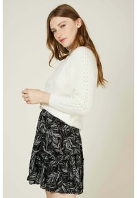 Pull en tricot blanc avec des détails en dentelle sur les manches, associé à une jupe noire présentant un motif argenté aux feuilles et un design en niveaux.