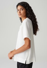 Haut blanc à manches courtes en tissu texturé, avec un décolleté arrondi et des fentes sur les côtés, associé à un pantalon noir.