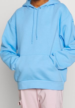 Person trägt hellblauen Hoodie mit Fronttasche und Kordeln, kombiniert mit hellrosa Cargohose.