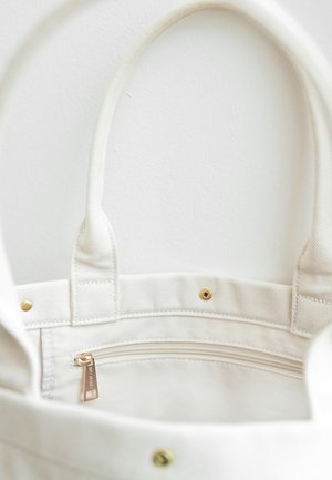 Witte canvas tas met twee stevige handvaten, voorzien van een ritsvak aan de binnenkant, verfraaid met gouden hardware en drukknoopsluitingen.
