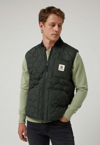 Chaleco acolchado de color verde oscuro con cuello alto, cierre de cremallera y dos bolsillos delanteros. Parche con el logo en el pecho, llevado sobre una camisa de manga larga de color verde claro.