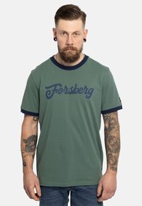 Camiseta verde con cuello redondo y mangas acanaladas en azul marino, que presenta un gran logo en azul marino de "Forsberg" en el pecho. Textura de tela suave.