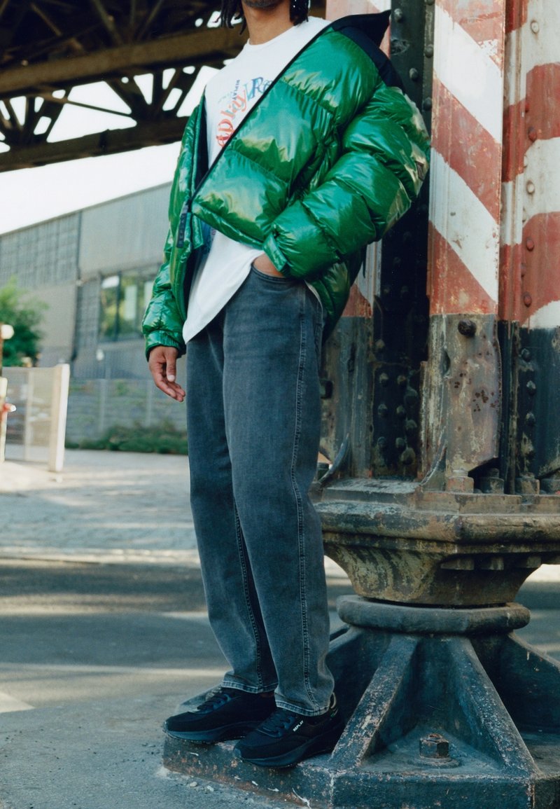 Personne portant une veste matelassée verte brillante, un t-shirt blanc graphique, un jean gris délavé et des baskets noires debout près d'un pilier industriel usé en extérieur.