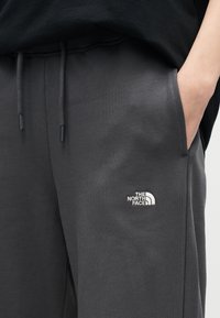 Κοντινό πλάνο στο χέρι ενός ατόμου στην τσέπη σκούρου γκρι φόρμας με το λογότυπο The North Face κεντημένο στον μηρό.