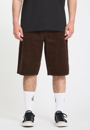 Personne portant un short en velours côtelé marron foncé, des chaussettes blanches mi-mollet avec un logo noir, des baskets basses noires et un t-shirt noir.