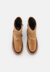 Zamszowe botki za kostkę w kolorze jasnobrązowym z podszyciem z sztucznego futra, z ozdobą w postaci złotego łańcuszka i czarną fakturowaną lamówką wokół podeszwy. Zapięcie na zamek błyskawiczny.