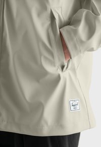Chaqueta de lluvia beige claro hecha de un material suave y waterproof, con un bolsillo lateral y un parche de logo blanco en el dobladillo inferior.