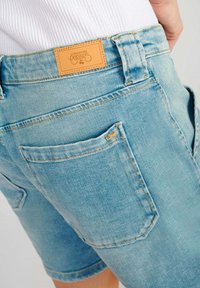 Shorts en denim bleu clair avec une étiquette en cuir à la taille arrière, deux poches arrière et des détails de couture visibles sur les coutures.