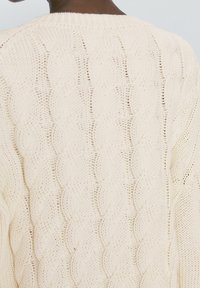 Pull over en câble beige clair avec un motif texturé, un col côtelé et une coupe décontractée, soulignant le design tissé.