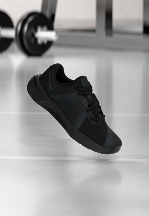 Sort atletiksko med tekstureret mesh-overdel, snøresystem og formet hæl. Har en sort gummiydersål og kontrasterende grønne logoaccenter.